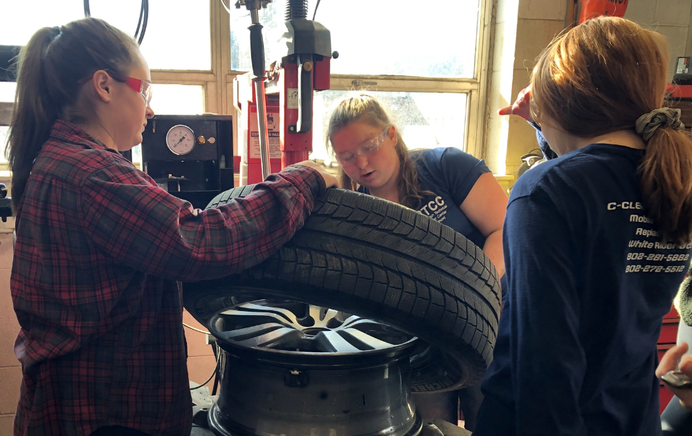 Female students demonstrate how to rotate a tire using a machine.