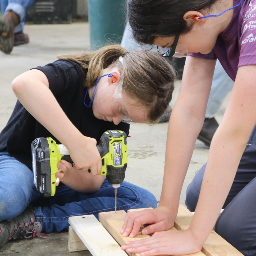 Rosie's Girls camper uses a drill to build a chair while a counselor in training assists.