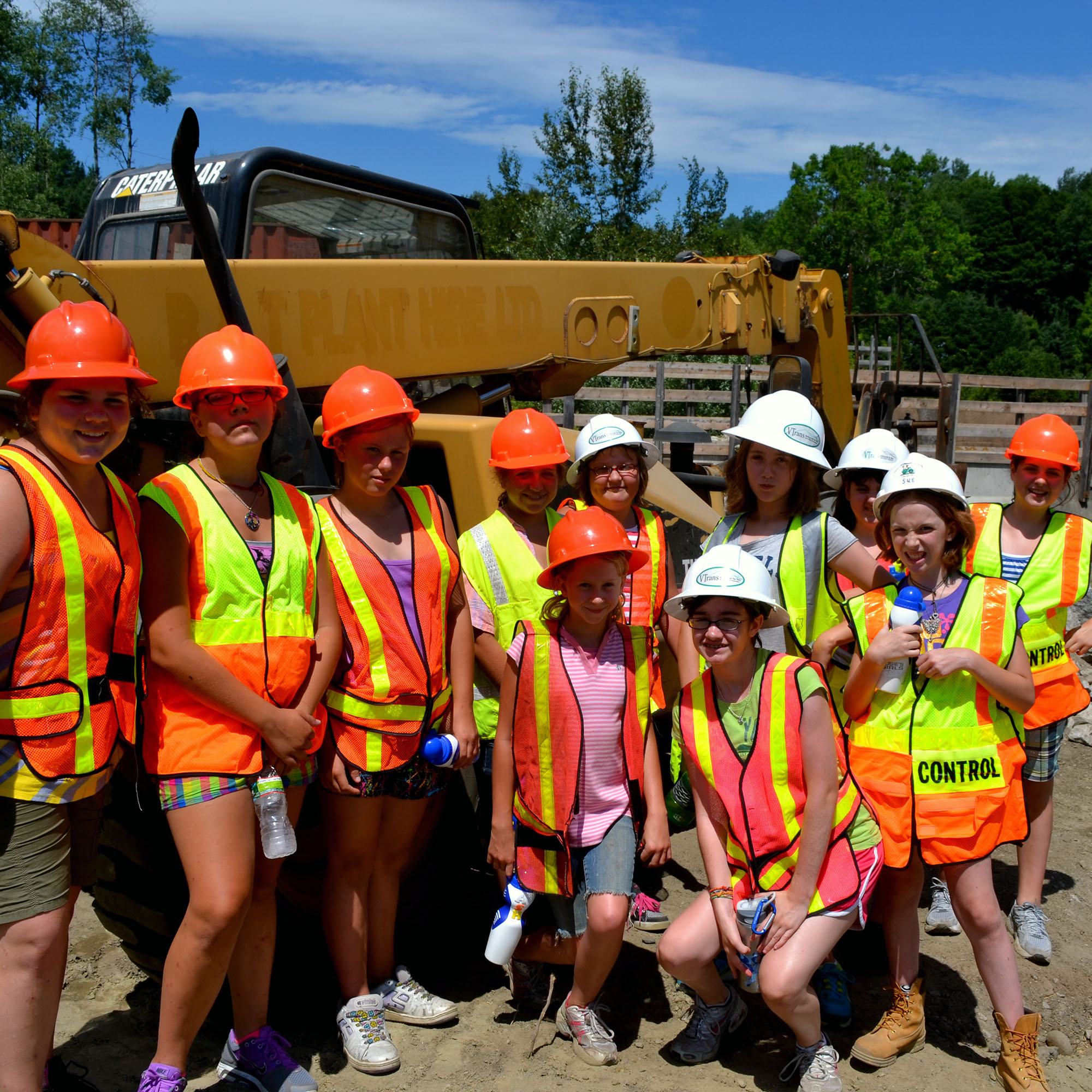 Rosie's Girls campers pose in front of a bridge wearing hard hats and high visibility vests. Vermont Works for Women's history dates back to 1987.