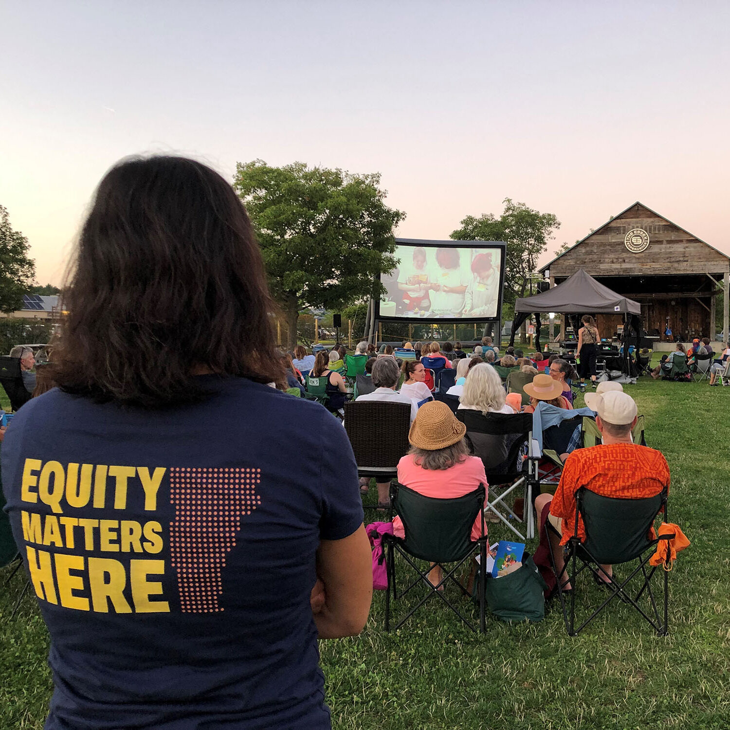 VWW staff member watches film at a LUNAFEST outdoor film screening. The back of her t-shirt reads Equity Matters Here. LUNAFEST is one of Vermont Works for Women's annual fundraisers.