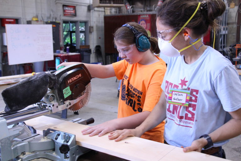 VWW staff member helps Rosie's Girls Camper use a saw to cut wood.