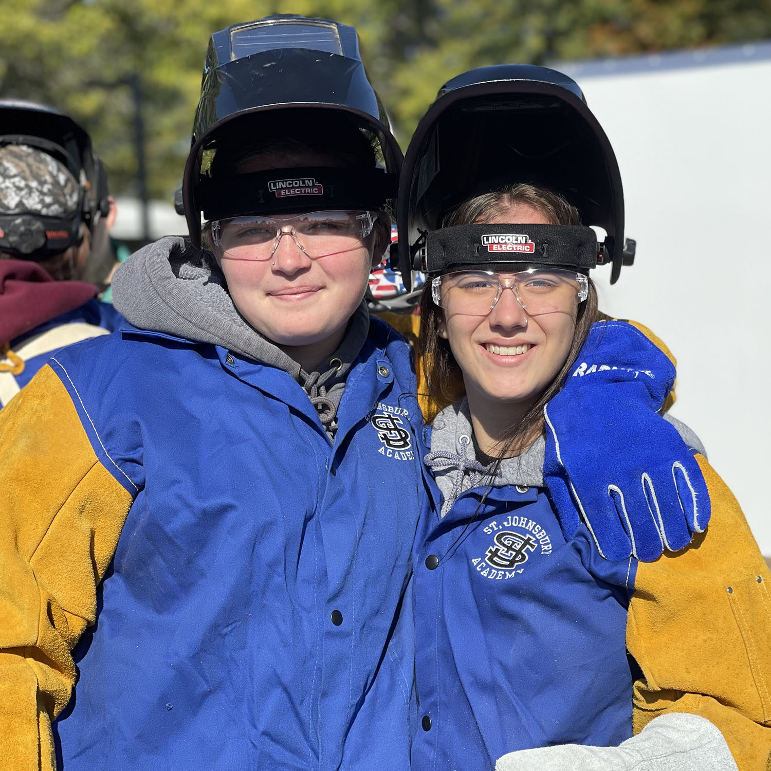 Two students smile while wearing welding jackets, gloves, and helmets.