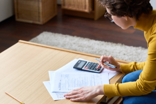 A woman is hunched over a coffee table, looking at papers and crunching numbers on a calculator.