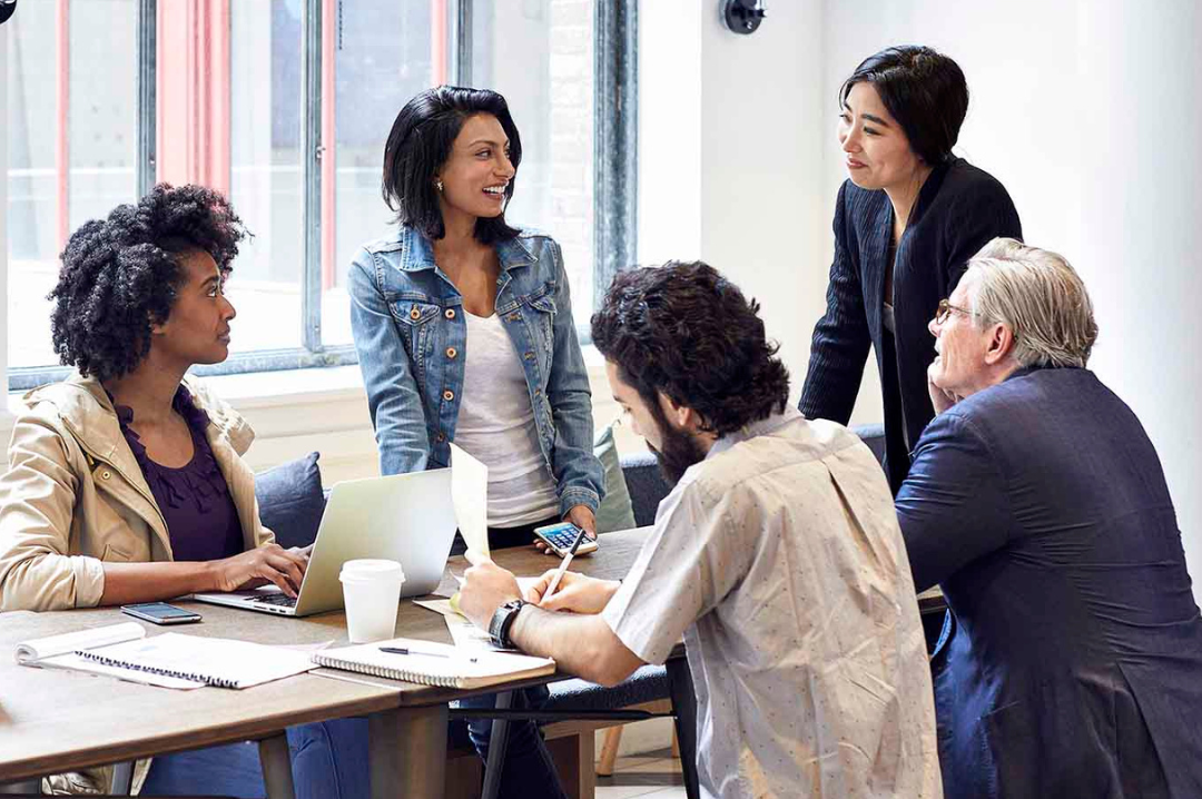 Five coworkers sit and stand around a table full of papers, computers, and beverages.