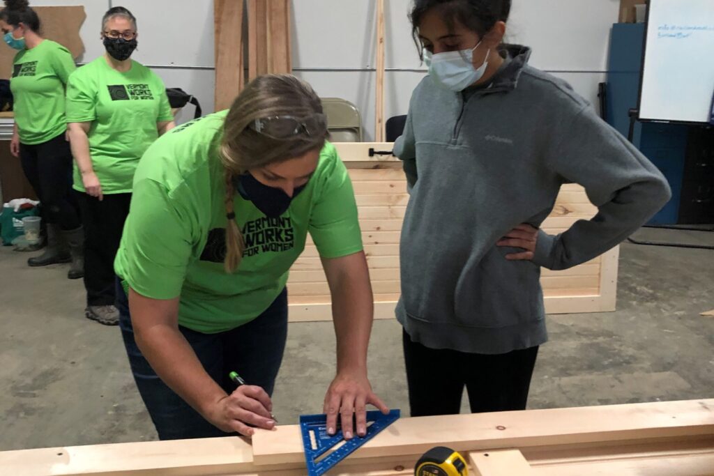 A woman wearing a Vermont Works for Women tshirts shows a high school aged individual how to measure a piece of wood.
