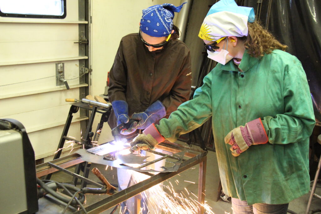 A Rosie's Girls Afterschool participant uses a plasma cutter to cut metal while a VWW staff member supervises.