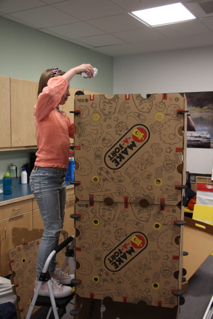 A Rosie's Girls Afterschool participant tests out their design, dropping the package from on top of a ladder through a cardboard fort.
