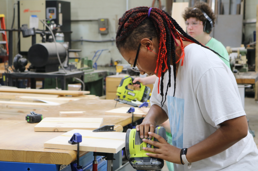 Rosie's Girls camper uses a sander on a piece of wood.