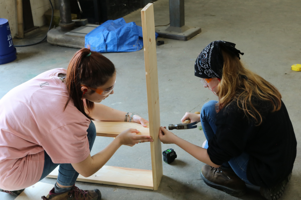 A CIT helps a Rosie's Girls camper as she hammers a nail while building a bookshelf.