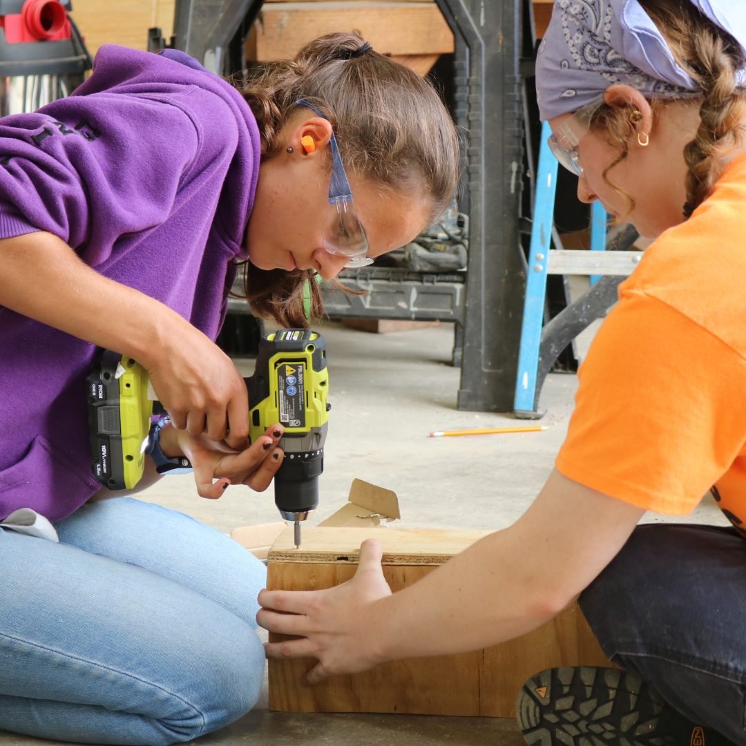 Rosie's Girls camper uses a drill to build a wooden project at camp with the help of an instructor
